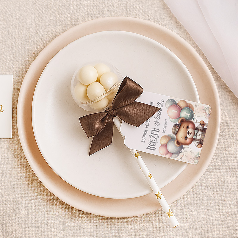 Białe cukierki w przezroczystym pojemniku spięte brązową kokardą, leżące na białym talerzyku z różowym brzegiem. Obok znajduje się karteczka z uroczym wzorem balonów i rysunkiem misia, zawierająca napis "Słodkie podziękowanie" w języku polskim oraz nienarzucający się słomka w białe gwiazdki.