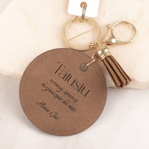 Brelok w kształcie okrągłej zawieszki z brązowego materiału, z napisem 'Tatusiu' oraz tekstem: 'Wymagaj zawsze bezpieczeństwa do nas', zakończony ciemniejszym frędzlem. Brelok zawieszony na metalowym kółku z dodatkowym chwytakiem.