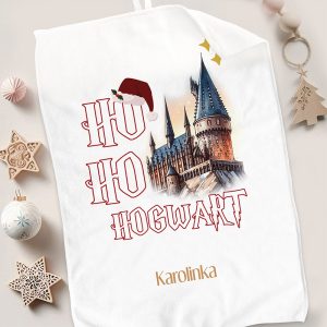 Biały ręcznik z nadrukiem przedstawiającym zamek, w stylu przypominającym Hogwarts, z napisem "Ho Ho Hogwart" i małą czerwoną czapką Mikołaja na górze. W tle dekoracje bożonarodzeniowe, takie jak gwiazdy i bombka.