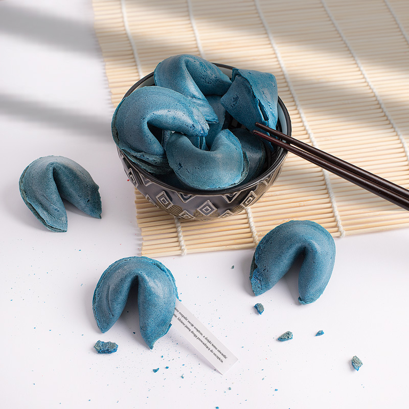 Miska z niebieskimi ciasteczkami fortuny, obok kilka pustych ciasteczek na tle bambusowej maty.