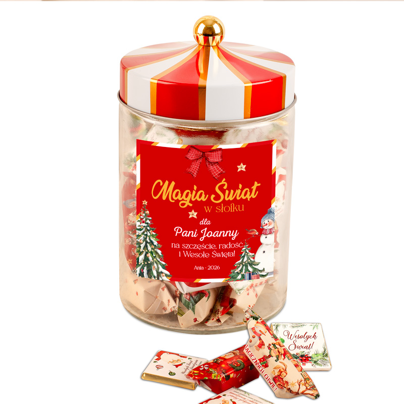 Szklany słoik z czerwonym wieczkiem, zawierający kolorowe karteczki z życzeniami bożonarodzeniowymi, z etykietą 'Magia Świat w słoiku dla Pani Joanny na szczęście, radość i Wesołe Święta'. Wokół słoika widać dekoracje świąteczne, a obok leżą małe czekoladki z życzeniami.