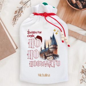 Biały worek z czerwonym sznurkiem, na którym znajduje się grafika przedstawiająca zamek oraz napis "Świąteczne ciepło HO HO HOGWARTU". Na worek nałożona jest czerwona czapka Mikołaja.