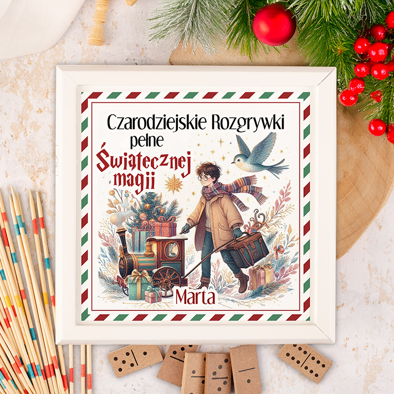 Ilustracja o tematyce świątecznej przedstawiająca młodego chłopca z torbą i wózkiem, w którym znajduje się mała lokomotywa oraz prezenty. W tle widać choinkę i lecącego ptaka. Tytuł książki "Czarodziejskie Rozgrywki pełne Świątecznej magii" znajduje się na górze ilustracji. Całość otoczona jest dekoracyjną ramką.