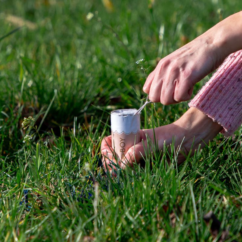 Osoba siedząca na trawie, trzymająca w ręku kubeczek lodów oraz łyżeczkę do nakładania, z nogą ułożoną obok na ziemi w jasnym swetrze.