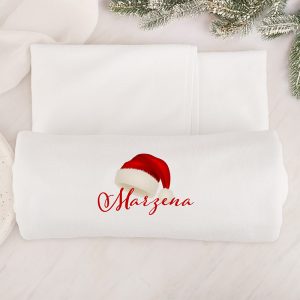 Biała kołdra z nadrukiem imienia 'Marzena' i czerwoną czapką na Mikołaja, leży na jasnym tle z gałązką choinki w górnym rogu