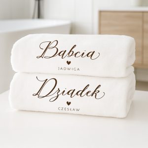 Dwie białe, miękkie ręczniki ułożone jeden na drugim, z napisami: 'Babcia Jadwiga' na górnym ręczniku i 'Dziadek Czesław' na dolnym, w eleganckim stylu czcionki z sercami w centralnej części.