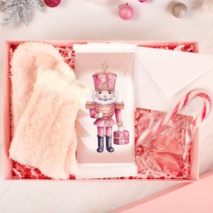 Różowy zestaw prezentowy zawierający miękkie skarpetki, ozdobne opakowanie z rysunkiem żołnierza z nutcracker, lizak w kształcie serca i kopertę, wszystkie otoczone kolorowymi ozdobami choinkowymi.