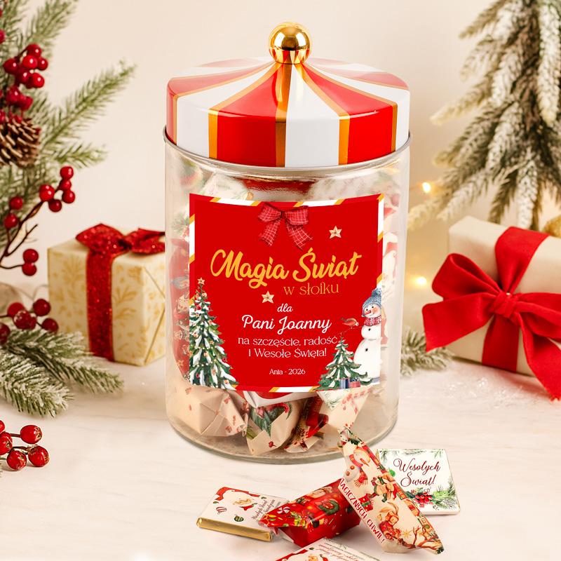 Słoik z napisem 'Magia Świąt w słoiku' przeznaczony dla Pani Joanny, udekorowany świątecznymi motywami, obok znajdują się zapakowane słodycze i prezent z czerwoną kokardą na tle choinki i świątecznych ozdób.