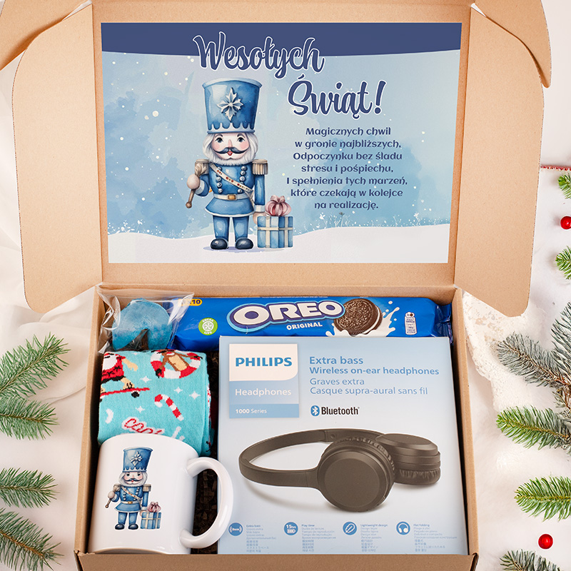 Otwarte pudełko prezentowe z niebieską kartką z postacią żołnierza świątecznego oraz napisem 'Wesołych Świąt!'. Wewnątrz znajdują się: paczka ciastek Oreo, słuchawki Philips, kolorowy kubek oraz otulacz. Tło z gałązkami świątecznymi.