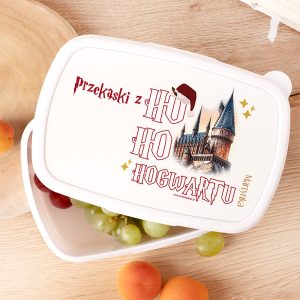 Biały pojemnik na jedzenie z napisem "przekąski z HO HO HOGWARTU" oraz rysunkiem zamku. W środku widoczne winogrona i brzoskwinie, a w tle drewniany stół.
