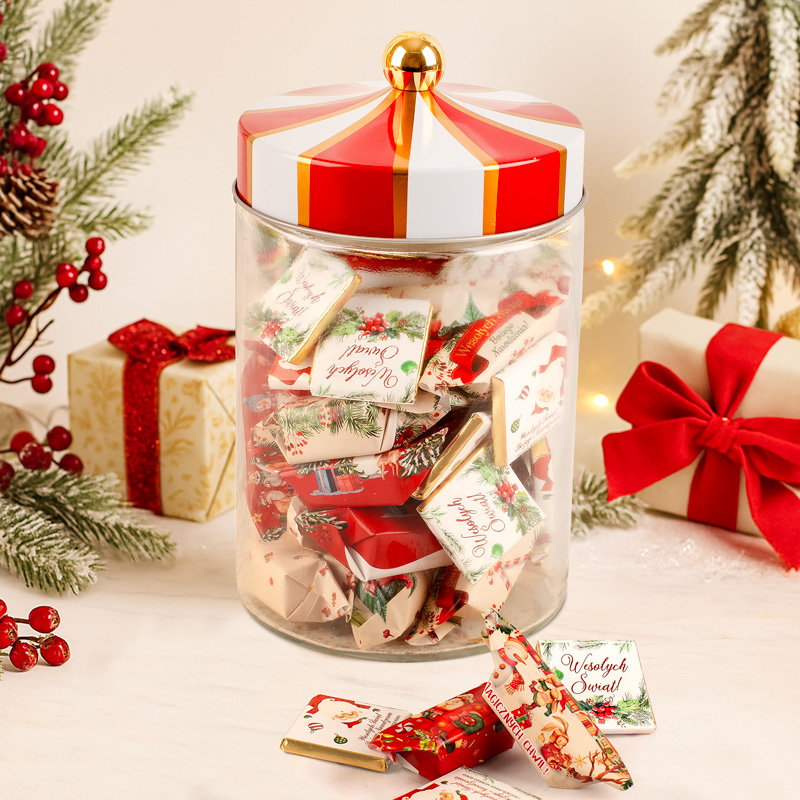 Transparent słoik z kolorową pokrywką w kształcie cyrkowego dachu, wypełniony słodkimi przekąskami z dekoracjami bożonarodzeniowymi, obok pakunek prezentu z czerwoną wstążką i choinka w tle.