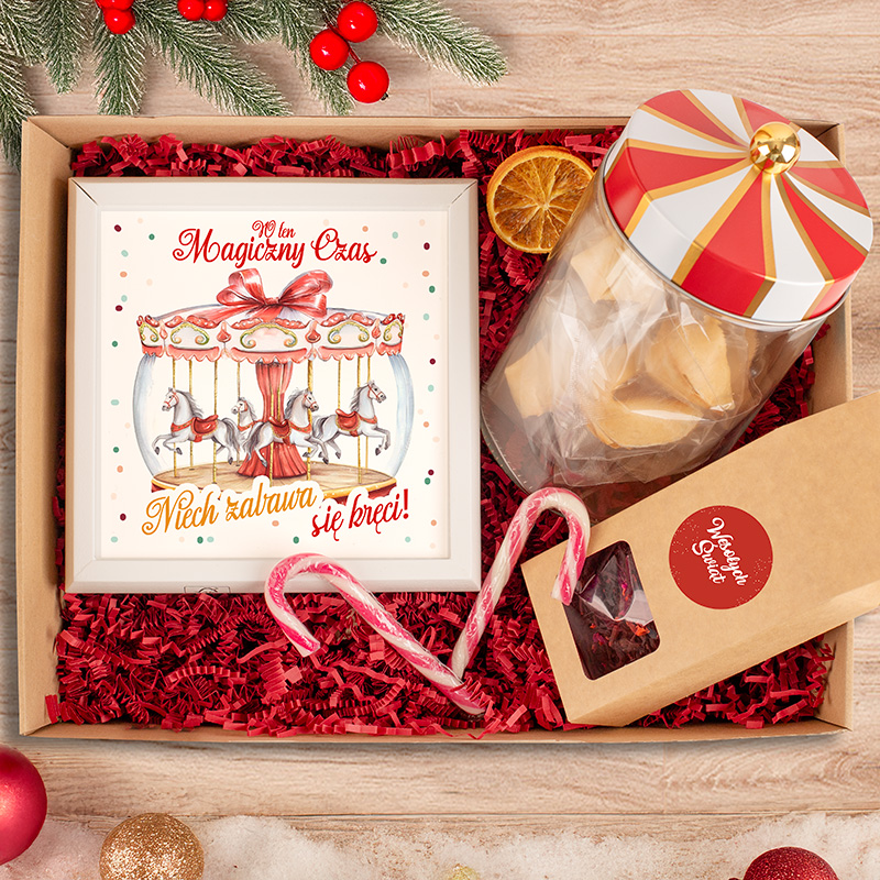 Stylowa paczka prezentowa zawierająca ramkę z ilustracją karuzeli, cukierki w słoiku z ozdobną pokrywką, czerwoną słomkę oraz pudełko z wypiekami, otoczona czerwonymi wstążkami i ozdobami świątecznymi.