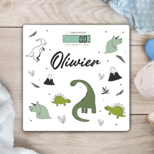 Waga elektroniczna ozdobiona ilustracjami dinozaurów oraz nazwą 'Olivier', wyświetlająca wartość 0.0 kg, położona na jasnym, drewnianym tle obok tekstyliów dziecięcych.