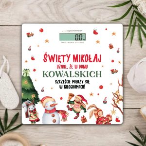 Waga elektroniczna z napisem 'Święty Mikołaj uznał, że w domu NAZWISKO szczęście mierzy się w kilogramach!', ozdobiona świątecznymi ilustracjami, takimi jak bałwanek, choinka, prezenty i postacie w świątecznych strojach, leży na drewnianym blacie otoczona dekoracjami bożonarodzeniowymi.