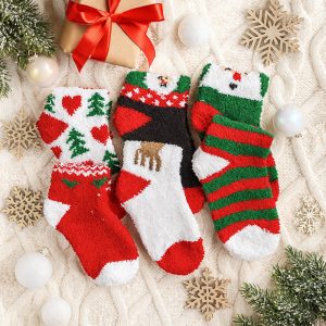 Trójka kolorowych, zimowych skarpet w świątecznych wzorach, umieszczonych na jasnym tle w otoczeniu dekoracji bożonarodzeniowych, w tym prezentu z czerwoną wstążką, śnieżynek i ozdób choinkowych.