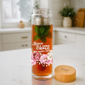 Przezroczysta butelka z napojem, w której unosi się świeża mięta, obok niej drewniana kapsułka z napisem 'RENATA'. Na butelce widnieje napis 'Babciu Dla Ciebie Ciepło Na Każdy Dzień'. W tle znajduje się jasna kuchnia.