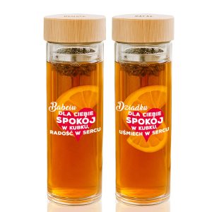 Dwa szklane kubki z herbatą, jeden z napisem dla Babci, a drugi dla Dziadka. Oba mają metalowe sitka i dekorację w postaci plasterka pomarańczy oraz tekst życzeń, w tym "spokój w kubku".