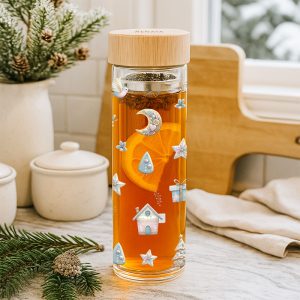 Przezroczysta szklanka z herbatą ozdobiona kolorowymi rysunkami, w środku plasterek pomarańczy, na blacie kuchennym w tle białe naczynia i gałązki sosny.