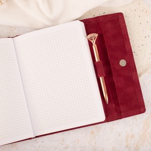 Otwarty notes z siatkowymi stronami w bordowej okładce, obok znajduje się różowo-złoty długopis.