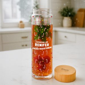 Przezroczysty dzbanek na herbatę z zielonymi liśćmi, czerwonymi owocami i dekoracjami w formie cukierków w paski, stojący na blacie kuchennym z drewnianą pokrywą z napisem 'RENATA'. W tle widoczna kuchnia z naturalnym światłem.