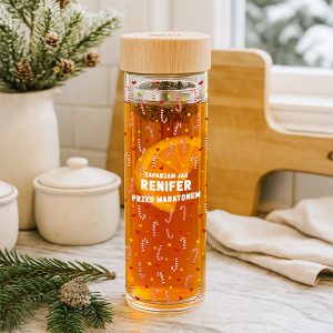 Przezroczysty dzbanek z herbatą, ozdobiony wzorem z cukierków, na tle kuchni z posegregowanym dekoracjami świątecznymi i ceramicznymi naczyniami.