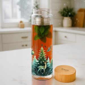Transparent szklanka z herbatą o pomarańczowej barwie, z liśćmi mięty na górze i kolorową ilustracją przedstawiającą choinkę oraz jelenia na dolnej części, umieszczona na białym blacie w kuchni.