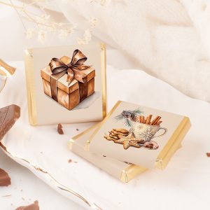 Dwa kawałki czekolady z ozdobnymi opakowaniami, jeden z wizerunkiem prezentu z kokardą, a drugi z kubkiem i ciasteczkami, leżące na talerzu obok kawałków czekolady i delikatnych dekoracji, takich jak gałązki i suszone kwiaty.