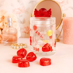 Słoik szklany ozdobiony kolorowymi motywami cukierków, z czerwonym wieczkiem w kształcie cukierka; obok leży kilka czerwonych cukierków na różowym tle, w tle widać perfumy i lusterko.