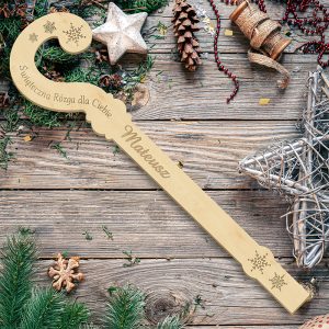 Drewniana świąteczna różdżka stylizowana w kształcie laski, z wygrawerowanym tekstem 'Świąteczna Różga dla Ciebie' i imieniem 'Mateusz', otoczona ozdobami bożonarodzeniowymi na drewnianym blacie.