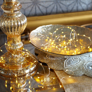 Złocona lampka na stole obok połyskującej talerza, na którym znajduje się świąteczne oznakowanie oraz drobne, świecące światełka.