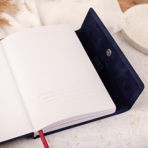 Otwarta notatnik z białymi kartkami, częściowo widoczna okładka w kolorze granatowym z materiału przypominającego zamsz, prezentująca miejsce na dane kontaktowe użytkownika na pierwszej stronie.