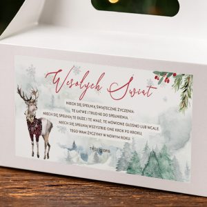Zbliżenie na białe pudełko z naklejką z napisem 'Wesołych Świąt' i ilustracją renifera w swetrze, otoczonym zimowym pejzażem oraz świątecznymi elementami takimi jak gałązki i płatki śniegu.
