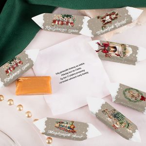 Na obrzeżach zdjęcia znajdują się kolorowe kartki z bożonarodzeniowymi motywami, opisane hasłami takimi jak "Rodzinnego czasu" i "Wesołych Świąt". Na środku leży biała koperta z wydrukowanym tekstem oraz kawałek słodyczy, a całość jest otoczona zielonym materiałem i złotymi perełkami.