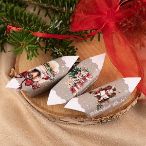 Dwa papierowe dekoracyjne elementy w kształcie pudełek, ozdobione malunkami bożonarodzeniowymi, leżące na kawałku drewna, obok gałązki iglastej i czerwonej wstążki. Na pudełkach znajdują się motywy związane z zimą i świętami, w tym kominek i postacie królików.