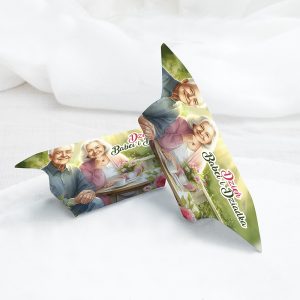 Grafika przedstawiająca kartkę z życzeniami z okazji Dnia Babci i Dziadka, z ilustracją uśmiechniętej pary seniorów, siedzących przy stole z filiżankami herbaty i kwiatami, na tle jasnych kolorów.