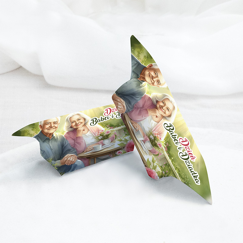 Grafika przedstawiająca kartkę z życzeniami z okazji Dnia Babci i Dziadka, z ilustracją uśmiechniętej pary seniorów, siedzących przy stole z filiżankami herbaty i kwiatami, na tle jasnych kolorów.