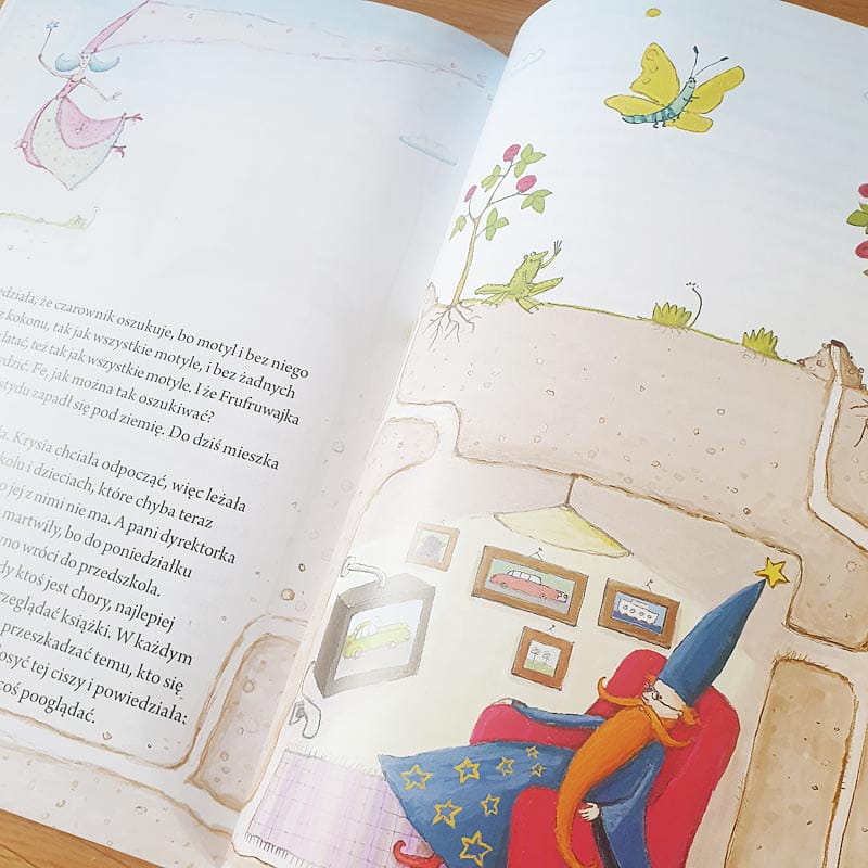 Ilustracja z książki dziecięcej przedstawiająca czarodzieja w niebieskim kapeluszu i czerwonym fotelu, otoczonego obrazkami na ścianie. Na stronie widać również motyle i rośliny, a w tle pojawia się sylwetka postaci w różowej sukni. Strony są jasne, z pastelowymi kolorami.