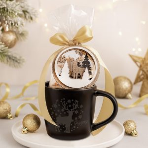 Czarny kubek z grawerowanym reniferem, ozdobiony złotą wstążką i przezroczystą folią, z ozdobą w kształcie choinki i renifera na górze, w otoczeniu złotych bombek i świątecznych dekoracji.