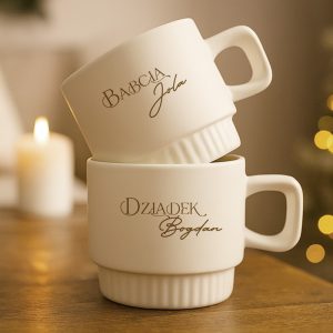 Dwie białe filiżanki ułożone jedna na drugiej, z napisem 'Babcia Jola' na górnej filiżance i 'Dziadek Bogdan' na dolnej, na tle rozmytego otoczenia świątecznego z lampkami.