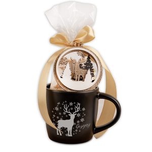 Czarny kubek z nadrukiem renifera i śnieżynek, ozdobiony złotą wstążką i przezroczystą folią, z zawieszką przedstawiającą leśny motyw z reniferem i drzewami.
