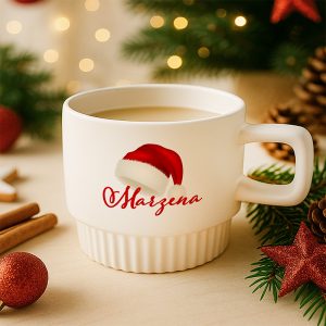 Biała filiżanka z logo 'Marzena', ozdobiona czerwoną czapką Mikołaja, wypełniona kawą, otoczona ozdobami świątecznymi, takimi jak gałązki iglaste, gwiazdki i bombki na tle rozmytego oświetlenia świątecznego.