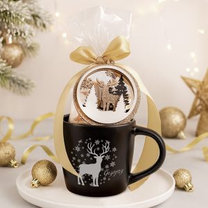 Czarny kubek z białym nadrukiem renifera i napisem "Gzygzzyz", obwiązany złotą wstążką. Na kubku umieszczony ozdobny okrągły element z przedstawieniem renifera i drzew. W tle dekoracje bożonarodzeniowe, złote bombki i choinkowe gałęzie.