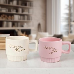 Dwa kubki na blacie: biały z napisem 'Dziadek Bogdan' oraz różowy z napisem 'Babcia Jola', za nimi nowoczesne wnętrze z półkami i kominkiem.