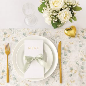 Elegancko nakryty stół z białym talerzem na obrusie w kwiatowy wzór, zawierający złoty widelczyk, nóż, szklankę i bukiet białych róż oraz serce w kolorze złotym obok talerza z menu przewiązanym zieloną wstążką.