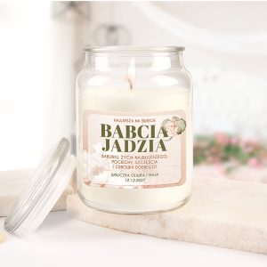 Słoik z świecą zapachową, na którym znajduje się etykieta z napisem 'Babcia Jadzia' oraz życzeniami z okazji urodzin. Słoik umieszczony na jasnym tle, z delikatnymi kwiatowymi wzorami w tle. Otwarta pokrywka obok słoika.