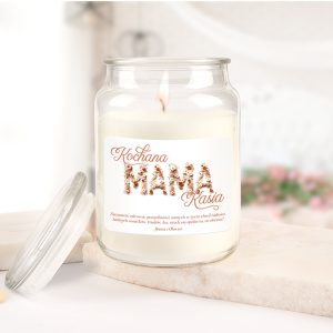 Słoik świecy zapachowej z etykietą, na której napisano 'Kochana MAMA Kasia', w otoczeniu jasnego tła i elementów dekoracyjnych. Świeca płonie, a wokół znajduje się subtelna aranżacja kwiatów.