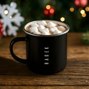 Czarny kubek z białymi piankami marshmallow na gorącej czekoladzie, z imieniem 'RADEK' na boku, ustawiony na drewnianym stole z rozmytym tłem świątecznych dekoracji.