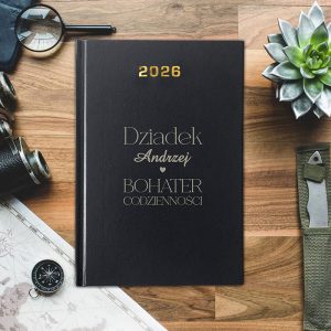 Czarna okładka zeszytu z napisem 'Dziadek Andrzej ♥ BOHATER CODZIENNOŚCI' oraz rokiem '2026', otoczona przyborami biurowymi, w tym kompasem i rośliną sukulentem, na drewnianym stole.