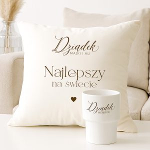 Jasna poduszka z napisem 'Dziadek MAJKI I ALI Najlepszy na świecie' oraz biała filiżanka z nazwiskiem 'Dziadek HENRYK', ozdobione serduszkiem; widok na stół i kanapę w tle.