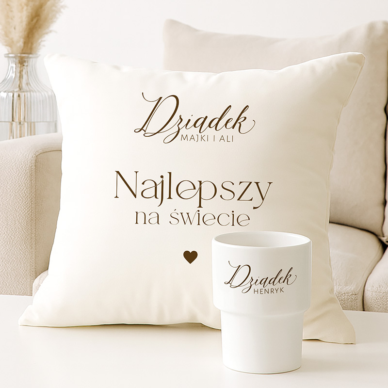 Jasna poduszka z napisem 'Dziadek MAJKI I ALI Najlepszy na świecie' oraz biała filiżanka z nazwiskiem 'Dziadek HENRYK', ozdobione serduszkiem; widok na stół i kanapę w tle.